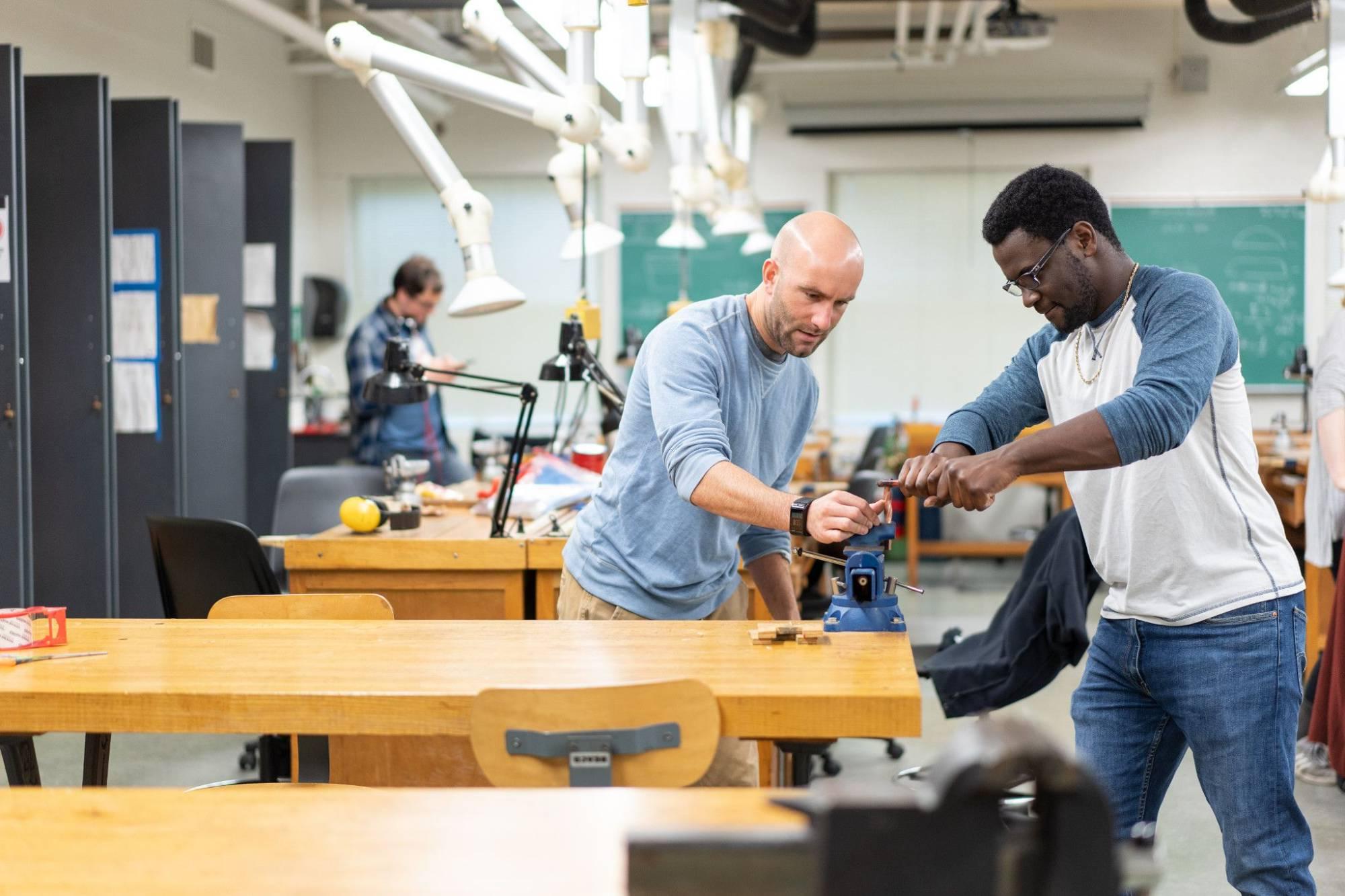 Image of a Black male student using equipment with the professor, Peter Antor, in a jewelry-making workshop. In the background there is more equipment and a young white male student.